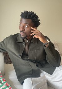 Rövid, göndör hajú és szakállas férfi sötétzöld kabátban és fehér nadrágban, beltéren ülve, gondolkodva oldalra néz.