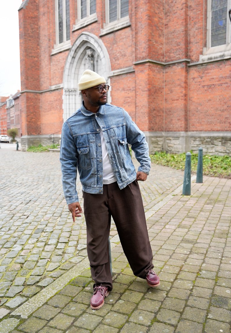 Homme portant un bonnet beige, des lunettes, une veste en denim, un pantalon marron et des chaussures bordeaux, debout sur une rue pavée près d'un bâtiment en briques.