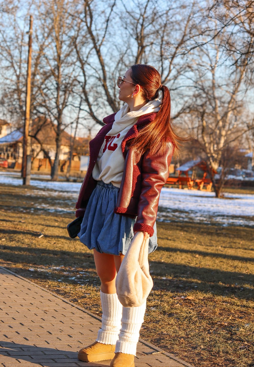 Bordo umetno usnjeno jopico s krznenim robom, bela pulover s kapuco, modra kavbojka krila, bele nogavice do kolen in svetlo rjave škornje, drži svetlo torbico.