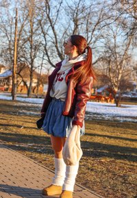 Bordo umetno usnjeno jopico s krznenim robom, bela pulover s kapuco, modra kavbojka krila, bele nogavice do kolen in svetlo rjave škornje, drži svetlo torbico.