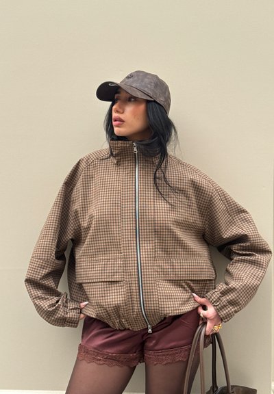 Mujer con chaqueta marrón a cuadros con cremallera, pantalones cortos granates con bordes de encaje, medias negras, gorra gris y sosteniendo un bolso marrón.