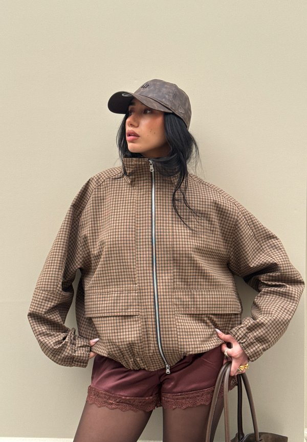 Femme portant une veste zippée à carreaux marron, un short bordeaux avec bordure en dentelle, des collants noirs, une casquette grise, et tenant un sac marron.