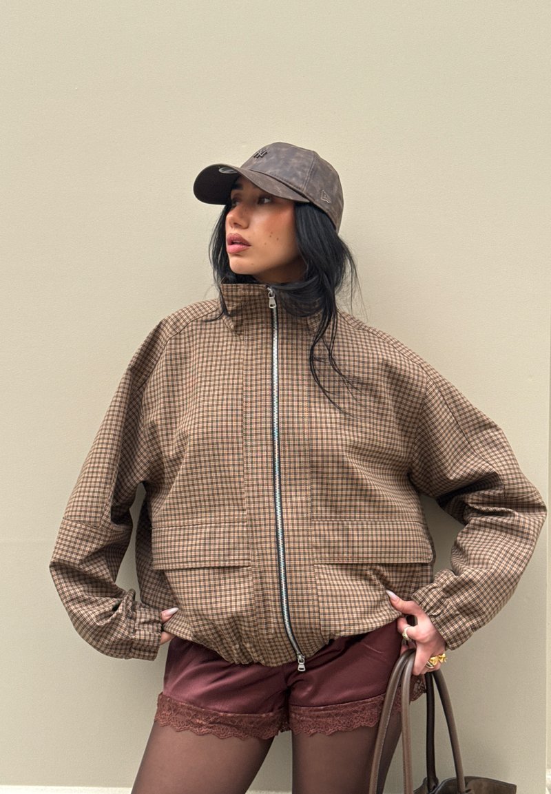 Vrouw draagt een bruine geruite jas met rits, bordeaux korte broek met kanten rand, zwarte panty's, een grijze pet en houdt een bruine tas vast.