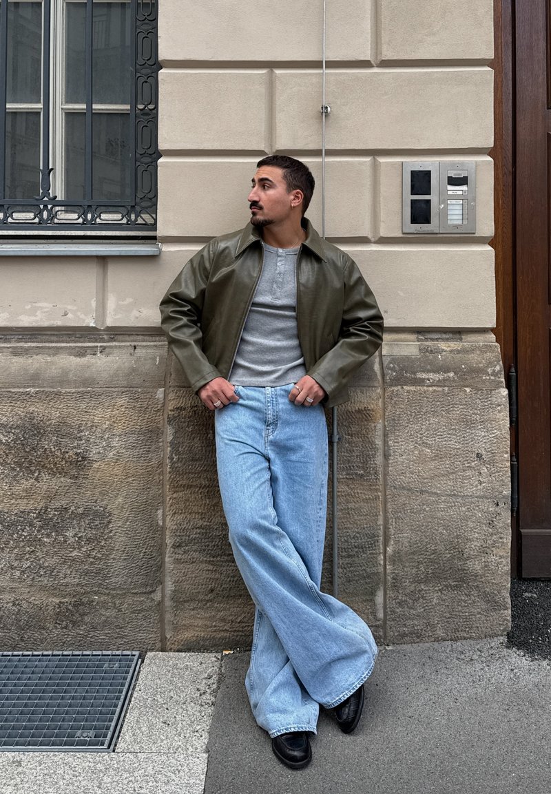 Homme aux cheveux courts et foncés et à la barbe appuyé contre un mur de pierre, portant une veste verte, une chemise grise, un jean large bleu et des chaussures noires.