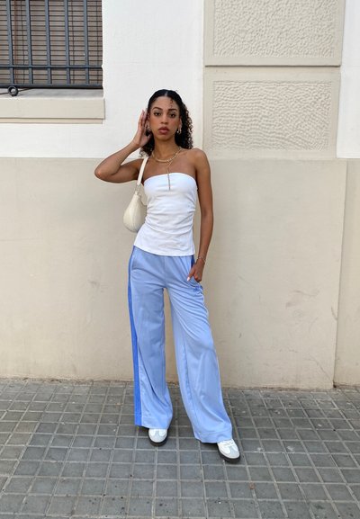 Top blanco sin tirantes; pantalones anchos de color azul claro con franjas laterales; zapatillas blancas; bolso pequeño blanco; collar de cadena dorada. Pared texturizada y pavimento.