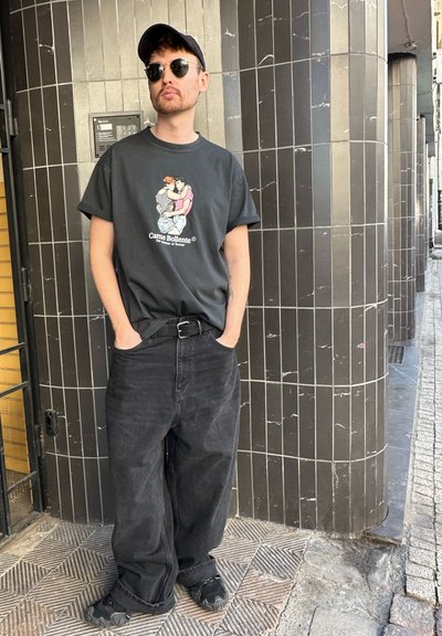 Camiseta negra con diseño gráfico, vaqueros negros holgados y zapatillas negras. Unas gafas de sol angulares y una gorra negra completan el look.