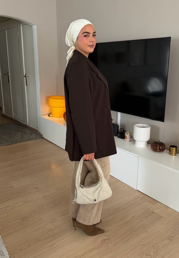Mulher usando lenço branco na cabeça e blazer escuro segurando uma mala bege, em pé numa sala de estar com uma TV e uma lâmpada atrás dela.