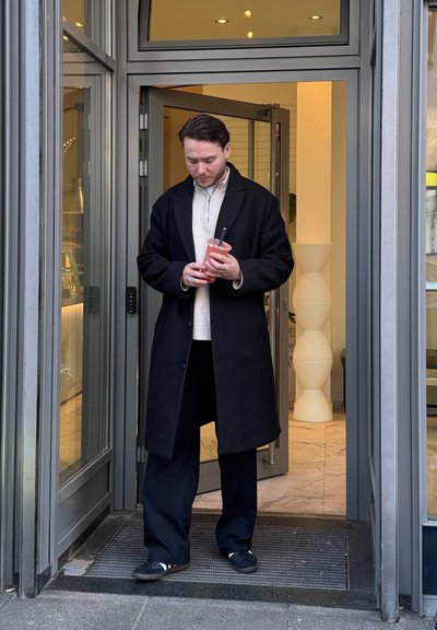 Abrigo negro sobre un suéter claro, sosteniendo una bebida rosa en un vaso transparente, de pie en una puerta con paneles de vidrio y suelo de baldosas.