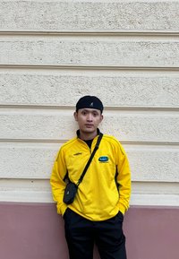 Chaqueta amarilla con cremallera y rayas horizontales, con un logo, combinada con pantalones negros y un pequeño bolso cruzado. Fondo de pared texturizada en tonos neutros.