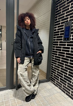 Chaqueta acolchada negra con capucha sobre un hoodie negro, combinada con pantalones cargo de camuflaje claros. Calzado negro. Pared de ladrillos texturizada de fondo.