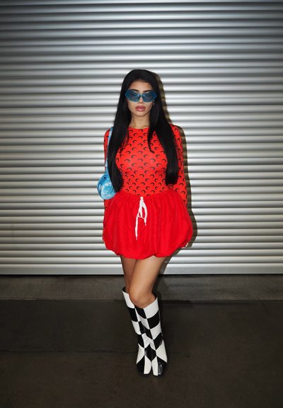 Bodysuit rojo de manga larga con patrones de media luna, combinado con una falda roja acampanada, un bolso azul y botas hasta la rodilla a cuadros en blanco y negro.