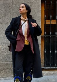 Zwarte jas, bordeauxblazer, crèmekleurige blouse, patroonshawl met franjes, donkere jeans met blauwe bloemborduursels. Tegen een textuurmuur.