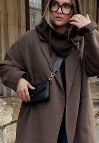 Cappotto in lana marrone oversize con cappuccio, abbinato a una sciarpa di lana lavorata a maglia marrone scuro. Una piccola borsa in pelle nera pende da una tracolla a fantasia.