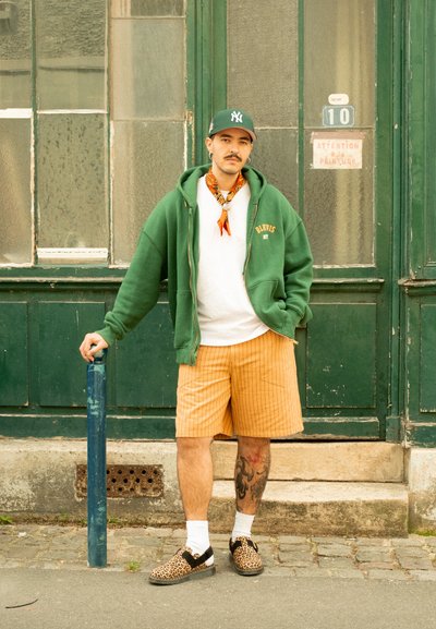 Sudadera verde con cremallera y texto 'BLKVIS', camiseta blanca, pantalones cortos a rayas naranjas y zapatos de estampado de leopardo. De pie con una mano apoyada en un poste azul.