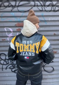 Μαύρο δερμάτινο τζάκετ με κίτρινες, λευκές και γκρι λεπτομέρειες. Διαθέτει τυπωμένο "TOMMY 1985" και "JEANS" στην πλάτη. Συνδυάζεται με καπέλο σε μπεζ απόχρωση.