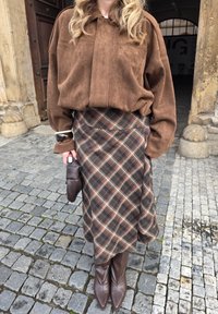 Pruun seemisnahast ülikure pikk särk, ruuduline pruun-beez seelik ja pruuni terava ninaga saapad. Käes hoidmas pruuni käekotti. Mudaäärega tänava taust.