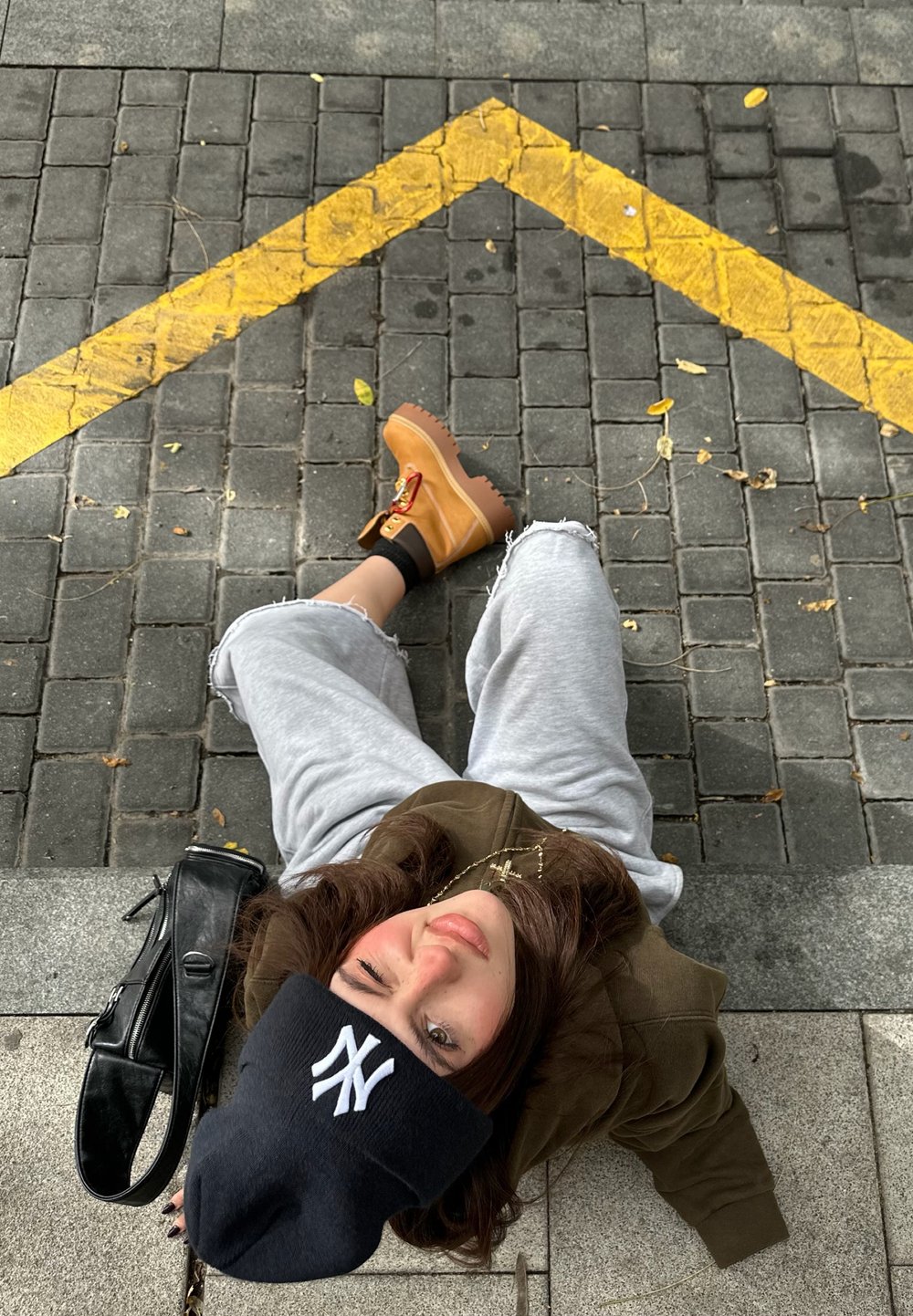 Personne assise sur une surface pavée, portant un sweat à capuche vert et un pantalon de survêtement gris, avec un bonnet noir, des bottes à plateforme jaunes et un sac noir.