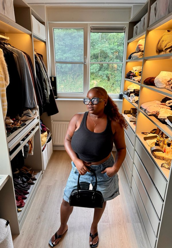 Vrouw die een zwart tanktopje en denimshorts draagt, houdt een zwarte handtas vast in een goed georganiseerde inloopkast met kleren en schoenen op planken.
