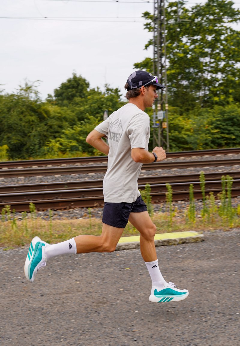 Homme portant une casquette noire, un t-shirt clair, un short noir et des chaussures de course blanches et turquoise, jogging près des voies ferrées et de la verdure.