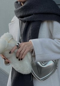 Λευκό εξωτερικό παλτό, σκούρο γκρι κασκόλ; κρατάει  μια αφράτη λευκή θήκη και μια λαμπερή ασημένια τσάντα σε σχήμα καρδιάς. Ορατά μαύρα νύχια και ένα μεταλλικό δαχτυλίδι.