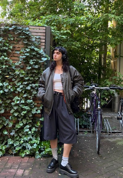 Bómbers verde sobre una camiseta blanca, pantalones cortos anchos negros, zapatos de plataformas negros, calcetines grises, con auriculares, rodeado de vegetación y bicicletas.