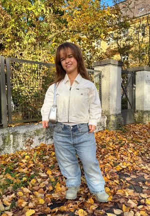 Chaqueta blanca corta con cremallera, vaqueros oversize azul claro con embellishments, y zuecos grises frente a un fondo de hojas de otoño.