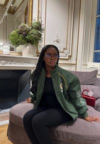 Mujer con cabello largo y negro y gafas, sentada en un sofá gris, vistiendo una chaqueta verde y ropa negra en una habitación con paredes blancas ornamentadas y una planta grande.