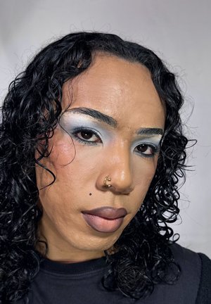 Primer plano de una persona con tono de piel medio, cabello rizado negro, sombra de ojos azul sutil y un piercing dorado en el septum. Labios nude llamativos con una pequeña marca de belleza.