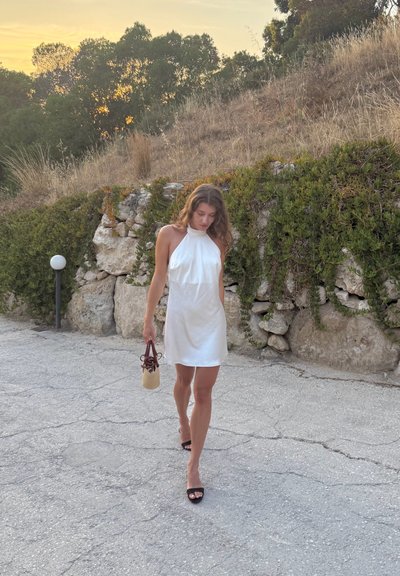 Vestido blanco sin mangas con un corte holgado, combinado con tacones negros. Sosteniendo un bolso beige con detalles marrones. El fondo presenta hierba y rocas.
