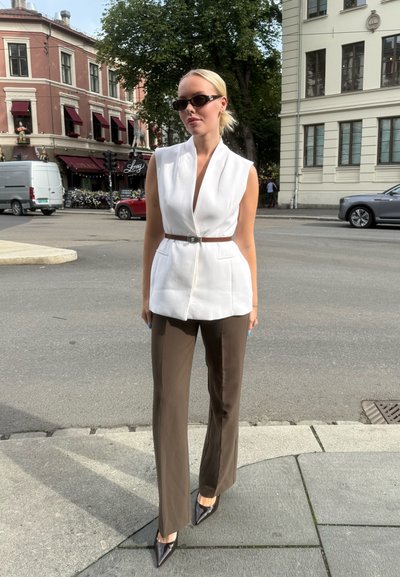 Mujer vistiendo un blazer blanco sin mangas con cinturón marrón, pantalones marrones y tacones negros de punta, de pie en la acera de la ciudad con edificios y coches cerca.