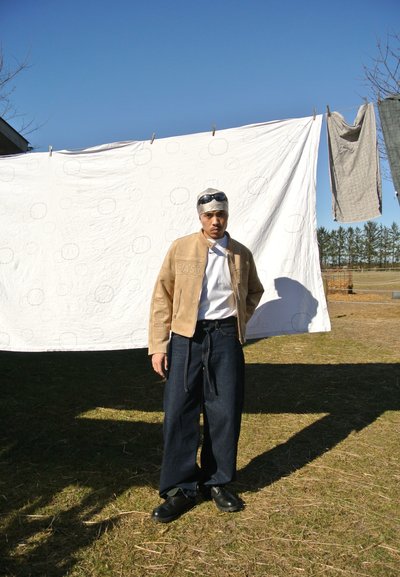 Chaqueta de cuero marrón con texto en relieve, camiseta blanca, pantalones de mezclilla oscuros y holgados, y zapatos negros. El fondo muestra ropa colgada en una cuerda.