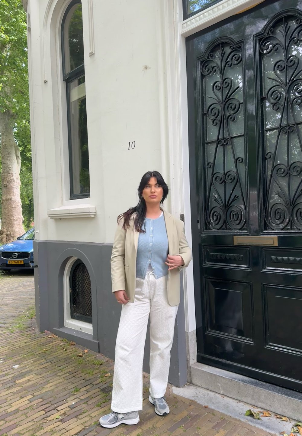 Light beige blazer over a blue button-up shirt, white textured wide-leg trousers, and grey sneakers. Ornate black door in the background.