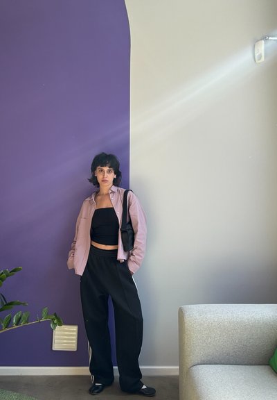 Camisa de cuadros con botones, top corto negro, pantalones de chándal negros con rayas blancas; de pie contra una pared morada y gris. Planta verde en primer plano.