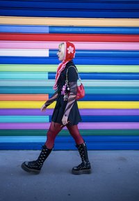 Modellen har på seg en svart minikjole med gjennomsiktige ermer, røde tights og svarte platåsko. En rød veske med myke nøkleringer. Fargerik stripete bakgrunn.