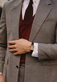 Casaco de fato cinza claro sobre um colete de malha bordô, camisa branca com abotoaduras e um relógio dourado com uma pulseira de couro preta.