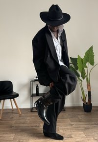 Cappello nero a tesa larga, blazer nero oversize su una camicia bianca, pantaloni neri su misura e stivali neri lucidi con dettagli testurizzati.