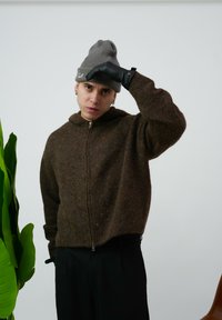 Brązowy sweter z kapturem i zamkiem, wykonany z teksturowanej tkaniny, zestawiony z szarym prążkowanym beanie oraz czarnymi rękawiczkami. Wnętrze z zieloną rośliną.