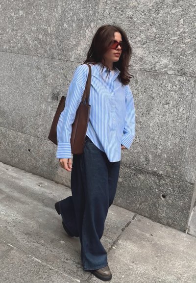 Camisa de botones a rayas azul y blanca, pantalones vaqueros oscuros y sueltos, bolsa de mano marrón y zapatos de plataforma marrones. El fondo presenta una pared texturizada de color gris.