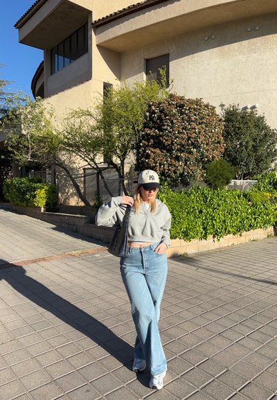 Mujer con sudadera gris corta, vaqueros azules, zapatillas blancas y gorra de béisbol caminando por una acera soleada cerca de un edificio con vegetación.