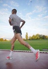 Corridore con tatuaggi che indossa pantaloni corti neri, maglietta grigia e scarpe da corsa grigie con strisce rosa, che corre su una pista bagnata con un campo verde e alberi.
