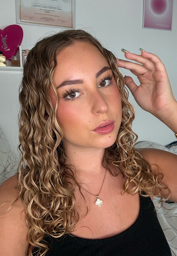 Jeune femme aux longs cheveux bouclés, portant un haut noir et un collier pendentif, touchant ses cheveux dans un environnement intérieur à la lumière douce.