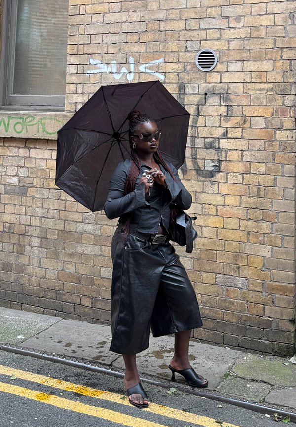 Pantalones culottes de cuero negro y chaqueta recortada, combinados con un paraguas negro y gafas de sol. Presenta accesorios únicos y un fondo de pared de ladrillo texturizado.