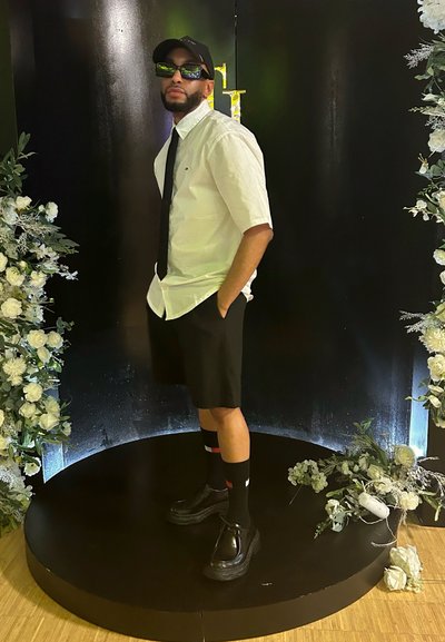 Camisa blanca de manga corta, corbata negra y pantalones cortos negros. Gafas de sol negras y gorra. Zapatos negros de suela gruesa y calcetines negros con un patrón en rojo y blanco.