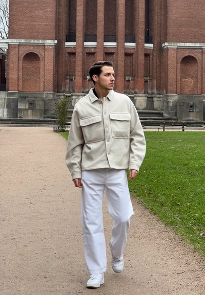 Chaqueta de felpa beige y de gran tamaño con dos bolsillos en el pecho, combinada con pantalones anchos blancos y zapatillas blancas. El fondo presenta una arquitectura de ladrillo rojo.