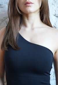 Hosszú barna hajú nő, fekete egyrészes vállpántos felsőt visel, egy világos, texturált fal előterében áll.