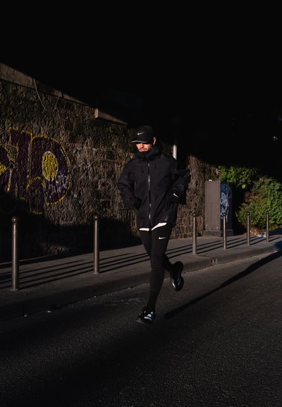 Chaqueta deportiva negra y leggings, zapatillas de correr, gorra oscura. El fondo muestra una pared de piedra con grafiti e iluminación callejera.
