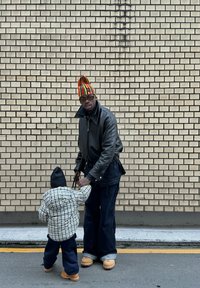 Man i svart läderjacka och färgglad randig hatt håller ett barns hand. Barnet har på sig en rutig pufferjacka och mörka byxor. Backdrop av tegelvägg.