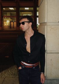 Camisa negra de manga larga parcialmente desabrochada, vaqueros azules, cinturón marrón, gafas de sol, de pie junto a un muro de piedra en un paisaje urbano nocturno.
