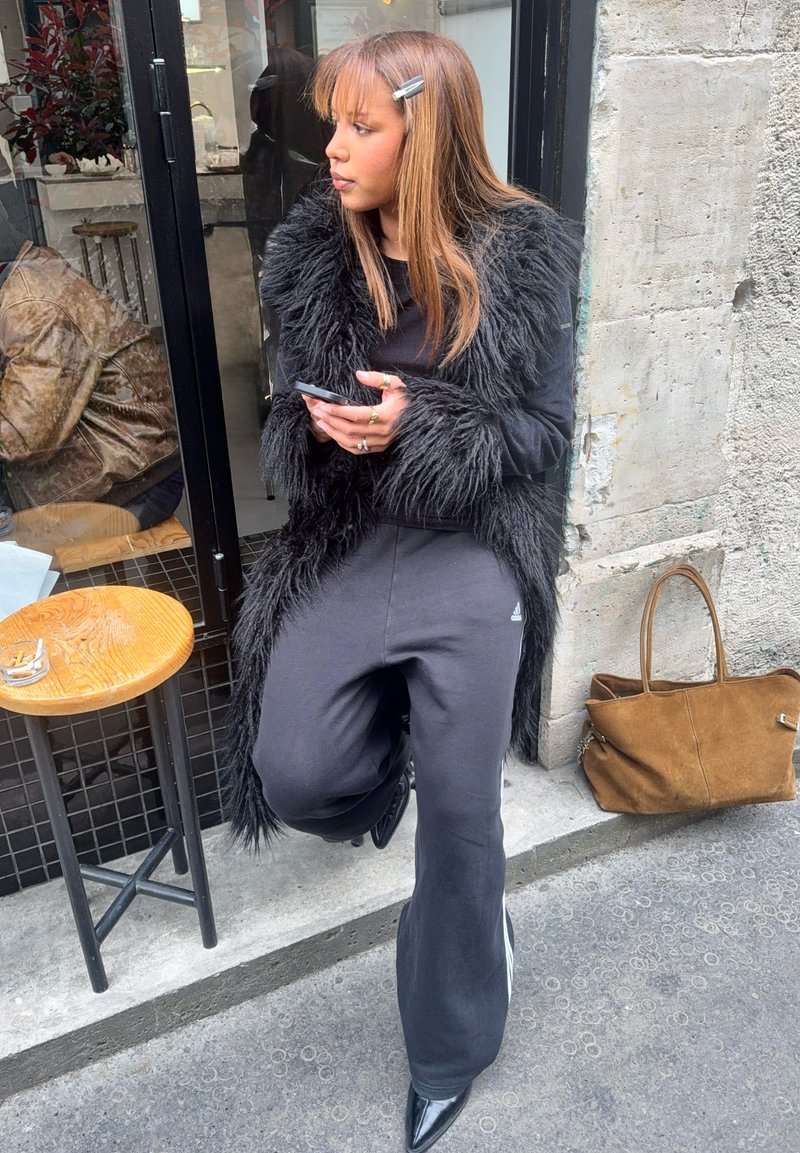 Femme en manteau noir en fourrure et pantalon noir debout devant un café, tenant un téléphone, avec un sac marron posé au sol contre un mur en béton.