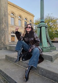 Hnedý kožený kabát s koženým golierom, čierne vysoké čižmy na opätku, modré rifľové džínsy a čierna kabelka, sediaci na kamenných schodoch pri stĺpe.